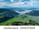 A sweeping panoramic view from the summit of Stanserhorn, featuring the tranquil Lake Lucerne surrounded by forested hills and green meadows at the mountains base. This scenic vista highlights the