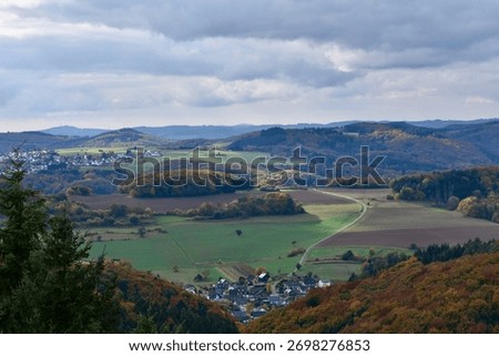 A sweeping valley view reveals patchwork fields, winding roads, and scattered villages nestled between forested hills under a brooding sky.