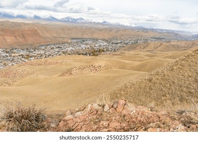 A sweeping view of a small town nestled in a valley surrounded by arid hills and mountains. Snow-capped peaks rise in the background, creating a stunning contrast with the golden terrain. - Powered by Shutterstock