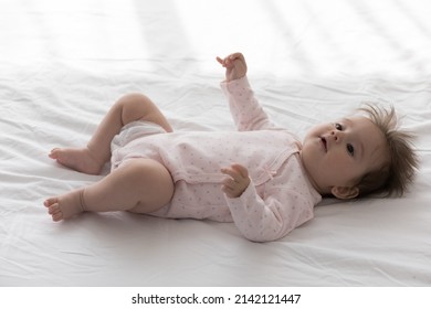 Sweet Adorable Few Month Baby Resting In Bed, Lying Back On Pale Linen Sheet, Looking Away With Interest, Curiosity. Infant Girl In Pink Bodysuit Relaxing On Soft Mattress. Childhood Concept