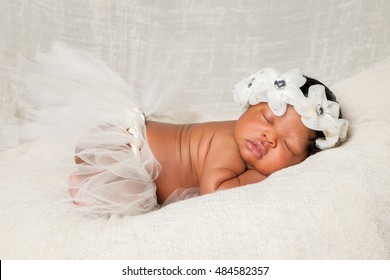 A sweet African American newborn sleeps quietly on an ivory blanket with a tutu and large floral headband.