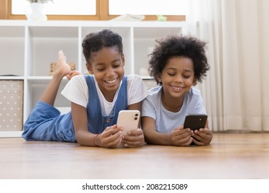 Sweet African sibling children relaxing on warm floor, enjoying leisure time with digital gadgets, using learning application on mobile phones, chatting on social media, watching online media content - Powered by Shutterstock