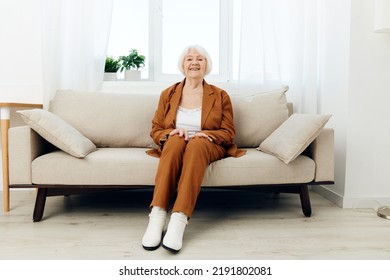 a sweet, amiable elderly woman in a brown suit is sitting on a beige wide sofa, smiling pleasantly while in the comfortable environment of her apartment - Shutterstock ID 2191802081