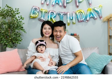 Sweet Asian Nuclear Family With A Lovely Baby Smiling At The Camera While Celebrating Birthday In A Modern Bright Living Room With Festive Decoration At Home