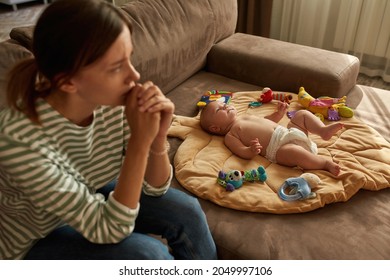 Sweet Baby Lying On Flat Pillow Placed On Sofa, Young Exhausted Mother Sitting Near, Looking Away. Single Mom Is Emotionally And Physically Overwhelmed By Her Parental Role. Parental Burnout Concept.