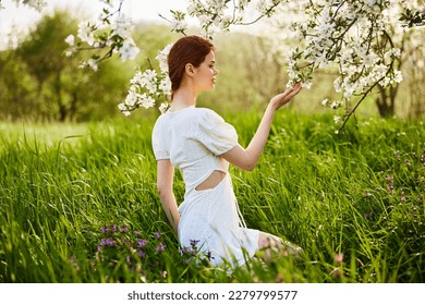 sweet, beautiful woman in a light short dress touches the branches of a flowering tree - Shutterstock ID 2279799577
