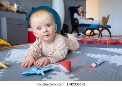 Sweet Blue Eyed Baby Playing On Floor Among Toys, Training To Crawl, Looking At Camera With Wide Eyes. Busy Mom Using Smartphone In Background. Childhood Or Staying At Home Concept