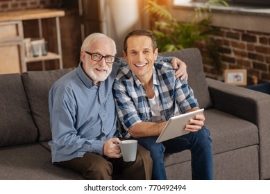 Sweet Bonding. Cheerful Elderly Man Sitting On The Sofa Next To His Adult Son, Hugging Him And Posing Together With Him For The Camera While Bonding To Each Other