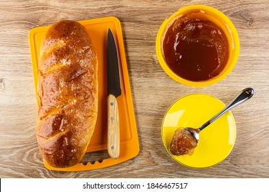 Sweet Bun, Kitchen Knife On Orange Cutting Board, Spoon With Apple Marmalade In Yellow Saucer, Plastic Jar With Apple Jam On Brown Wooden Table. Top View