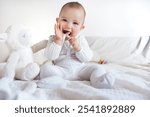 A sweet and candid image of a baby sitting on a bed, playfully biting a ribbon while holding a soft, plush stuffed animal. The baby, dressed in cozy, neutral-toned clothes, radiates joy and innocence.