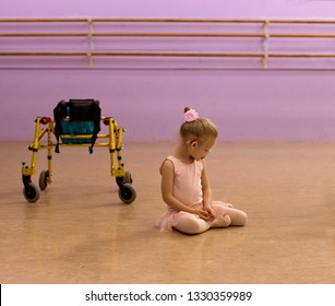 Sweet Caucasian Little Girl Wearing Hearing Aid, Glasses, A Bun And Pink Ballet Leotard Sits Next To Her Gold Walker Mobility Aid In Ballet Studio 