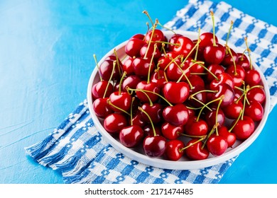 Sweet Cherries On A White Plate. Ripe Cherries And A White-blue Napkin On A Blue Background. Copy Space