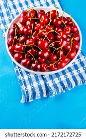 Sweet Cherries On A White Plate. Ripe Cherries And A White-blue Napkin On A Blue Background. Copy Space