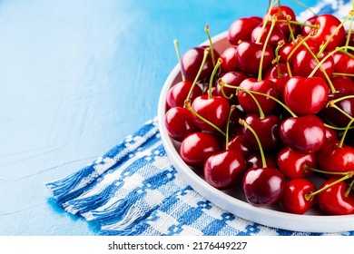 Sweet Cherries On A White Plate. Ripe Cherries And A White-blue Napkin On A Blue Background. Copy Space