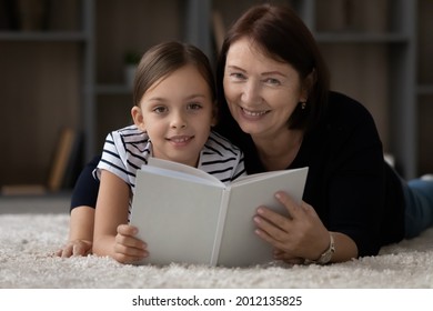 Sweet Child Reading Paper Book With Granny. Girl And Grandmother Resting On Heating Floor At Cozy Home, Looking Ant Camera, Smiling, Holding Textbook With Blank Cover. Headshot Portrait