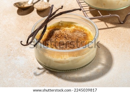 Sweet creme brulee dessert. Portioned glass jars with homemade creme brulee with caramelized sugar crust topping, on beige colored background with sugar in spoon and vanilla copy space