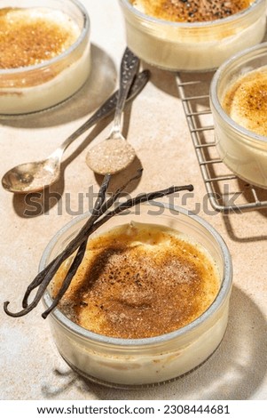 Sweet creme brulee dessert. Portioned glass jars with homemade creme brulee with caramelized sugar crust topping, on beige colored background with sugar in spoon and vanilla copy space