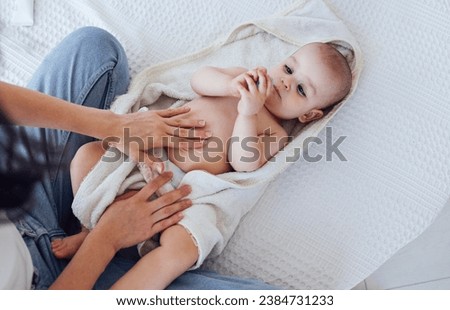Sweet cute baby on the bed next to his mom and plays with tube of cream. Young mother smears cream on her child. Light color bedroom interior