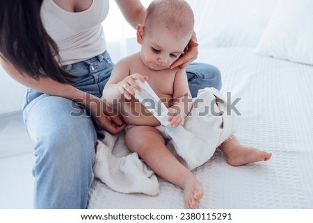 Sweet cute baby sits on the bed next to his mom and plays with tube of cream. Young mother smears cream on her child cheeks. Light color bedroom interior.