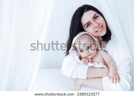 Sweet cute baby sits on the bed next to his mom and plays after bath. Light color bedroom interior