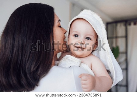 Sweet cute baby sits on the bed next to his mom and plays after bath. Light color bedroom interior