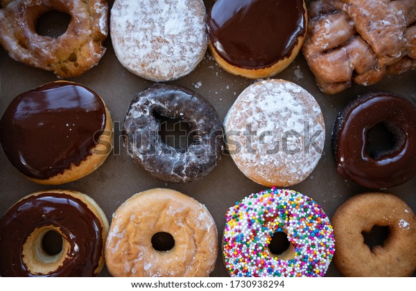 Sweet dozen of donuts in paper box. Picture\
of donut box with one dozen assorted fresh baked donuts- chocolate\
glazed, sprinkles, Boston cream, jelly, sour cream glazed, apple\
fritter and traditional.