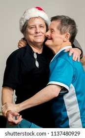 A Sweet, Elderly Man With Downs Syndrome Hugs And Dances With His Older Sister On Christmas Day.  She Is Wearing A Santa Hat And Trying Not To Laugh.  He Is Ecstatic And Has A Big Smile.  