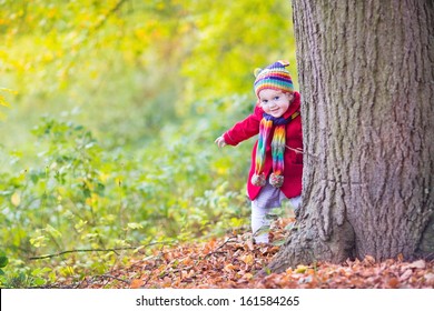 Sweet funny baby girl in a red coat and colorful hat and knitted scarf hiding behind a big old tree in a beautiful autumn park with yellow leaves