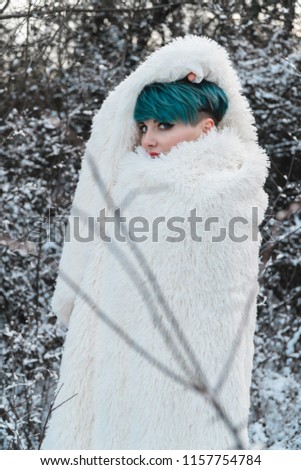 A sweet girl wrapped her head in a white fur coat, hiding from the cold in a winter snow-covered park