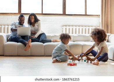 Sweet Home. Happy Loving Black Family Of Four Gather Together On Weekend, Young African Mom And Dad Relaxing On Sofa With Laptop And Two Kids Daughter And Son Having Fun Playing Toys On Heated Floor