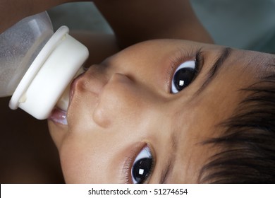 Sweet Indian baby with big eyes lying in parent's arms and being fed with baby bottle.