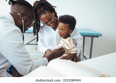 A Sweet Little African Boy Sits Up On The Exam Table At The Doctors, As His Male Doctor Listens To His Chest With His Stethoscope. The The Little Boy Is Dressed Causally And Is Giggling At The Doctor.