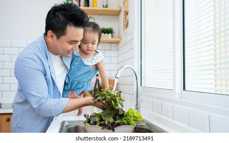 Sweet Little Asian Girl Helping Dad To Prepare Dinner, Making Salad From Fresh Vegetables. African American Young Father Teaching Kid To Cook In Home Kitchen At Table With Organic Food Ingredients.