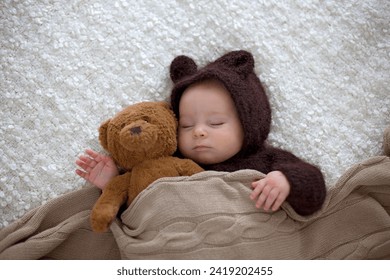 Sweet little baby boy, dressed in handmade knitted brown soft teddy bear overall, sleeping cozy at home in sunny bedroom with lots of teddy bears around him - Powered by Shutterstock