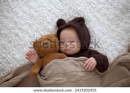 Sweet little baby boy, dressed in handmade knitted brown soft teddy bear overall, sleeping cozy at home in sunny bedroom with lots of teddy bears around him
