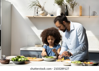 Sweet Little Black Girl Helping Dad To Prepare Dinner, Making Salad From Fresh Vegetables. African American Young Father Teaching Kid To Cook In Home Kitchen At Table With Organic Food Ingredients