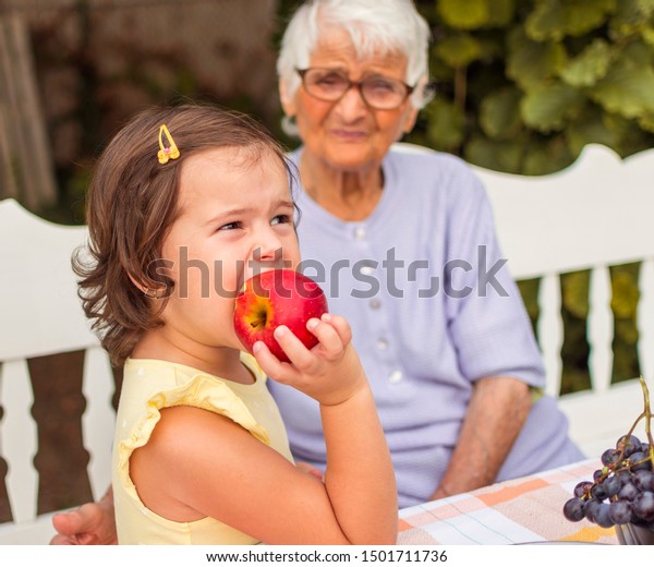 Sweet little girl eating an\
apple and spends time with her granny. Granny sit and enjoy to\
watching her heiress. Granny and granddaughter sitting at the table\
outdoor.