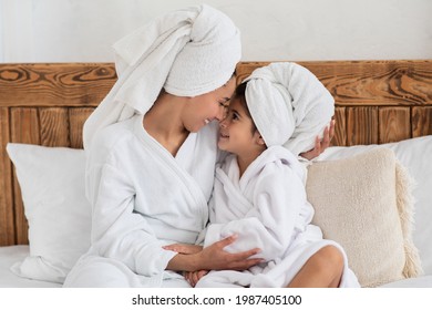 Sweet little girl having beauty day with her mother at home or at spa hotel, wearing white bathrobes, wrapping towels all over their heads, sitting on bed and cuddling, enjoying time together - Powered by Shutterstock