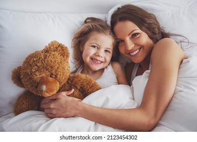 Sweet little girl and her beautiful young mother are looking at camera and smiling while lying with a teddy bear in bed at home - Powered by Shutterstock