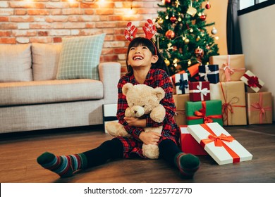 Sweet Little Girl Hugging Teddy Bear Looking Up To Sky Smiling Sitting On Wooden Floor. Attractive Childhood Received Gift On Christmas Sale At Home. Kid With Reindeer Satisfied With Present.