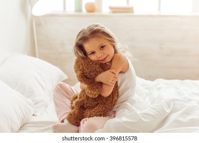 Sweet little girl is hugging a teddy bear, looking at camera and smiling while sitting on her bed at home