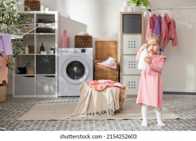 A Sweet Little Girl Plays In The Laundry Room Bathroom Next To The Laundry Basket With Clothes. Child Holds Bottle For Liquid, Soap Opens Cap Looks In, Curiosity Learning About The World.