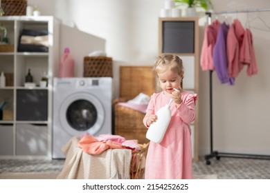 A Sweet Little Girl Plays In The Laundry Room Bathroom Next To The Laundry Basket With Clothes. Child Holds Bottle For Liquid, Soap Opens Cap Looks In, Curiosity Learning About The World.