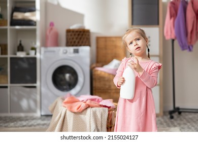 A Sweet Little Girl Plays In The Laundry Room Bathroom Next To The Laundry Basket With Clothes. Child Holds Bottle For Liquid, Soap Opens Cap Looks In, Curiosity Learning About The World.