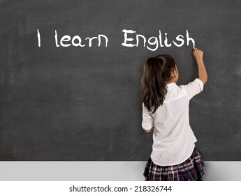 Sweet Little Hispanic Girl At School Lesson With Ponytail And Uniform Writing With Chalk On Classroom Blackboard In English Learning , Wisdom And Successful Education Concept 