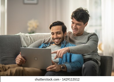 Sweet Male Queer Couple Spend Time at Home. They are Lying Down on Sofa and Use Laptop. They Browse Online. Partner's Hand is Around His Lover with Other Pointing on Screen. Room Has Modern Interior.