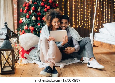 Sweet memories. Joyful black couple looking photos or video on digital tablet, sitting near Christmas tree at home, free space - Powered by Shutterstock