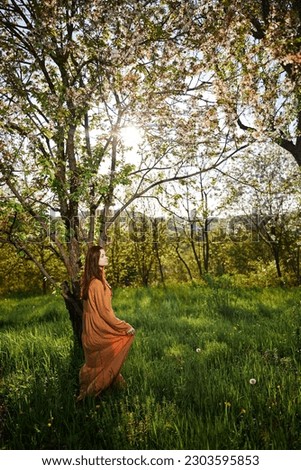 a sweet, modest, attractive woman with long red hair stands in the countryside near a flowering tree and lifts the hem of her dress up