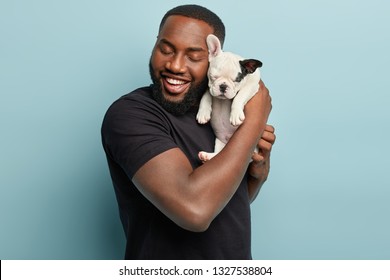 Sweet Moments With Domestic Animals. Portrait Of Satisfied Dark Skinned Man Holds Cute Small French Dog Puppy Closely To Face, Enjoys Good Time With Loyal Pet, Wears Casual Black T Shirt, Pose Indoor