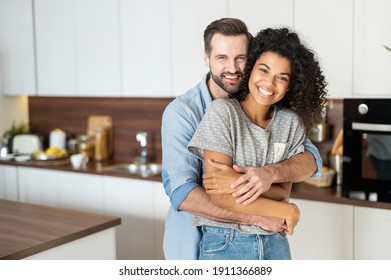 Sweet multi-ethnic couple in love stands in embraces in the kitchen. Cheerful an African woman and a caucasian guy look at the camera and smile toothy - Shutterstock ID 1911366889
